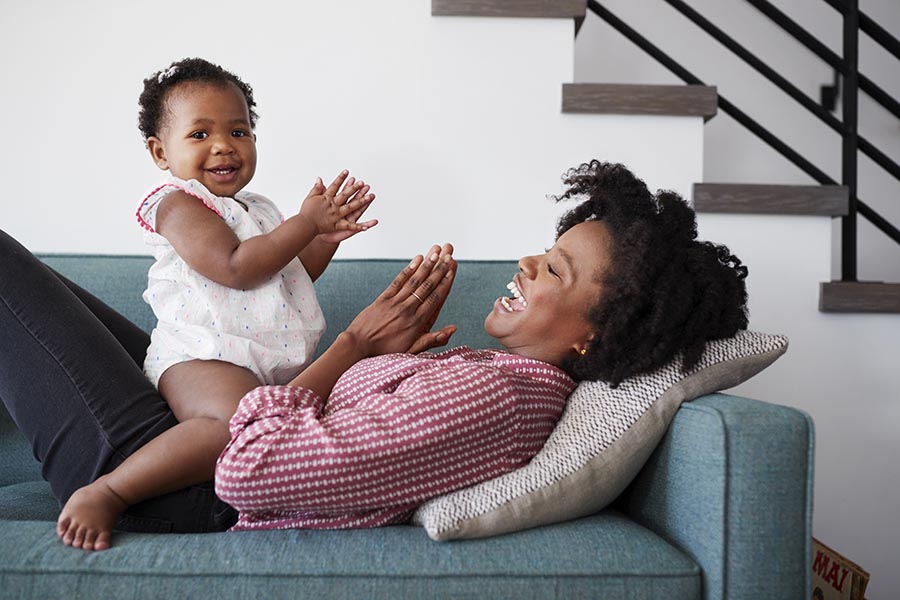 Personal Insurance - Mom and Baby Play Pat-a-Cake on Their Sofa, Baby on Mom's Lap, Both Smiling